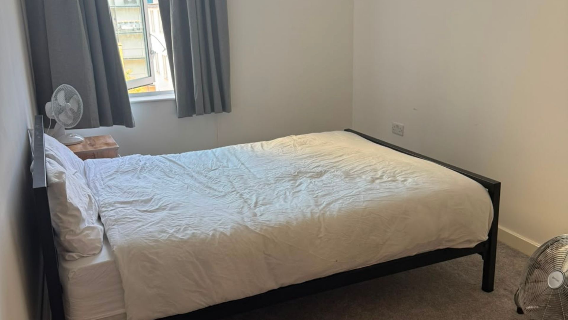 Un lit simple avec une couette blanche, une table de chevet, un ventilateur sur pied et une fenêtre avec des rideaux.