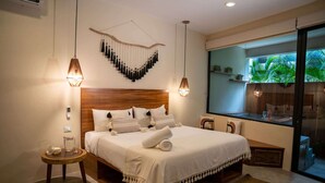 A bedroom with a wooden headboard, a large bed with white bedding, two bedside tables, a small round table, and a large window with a view of greenery. - Xperience Tulum 25 (Tulum)
