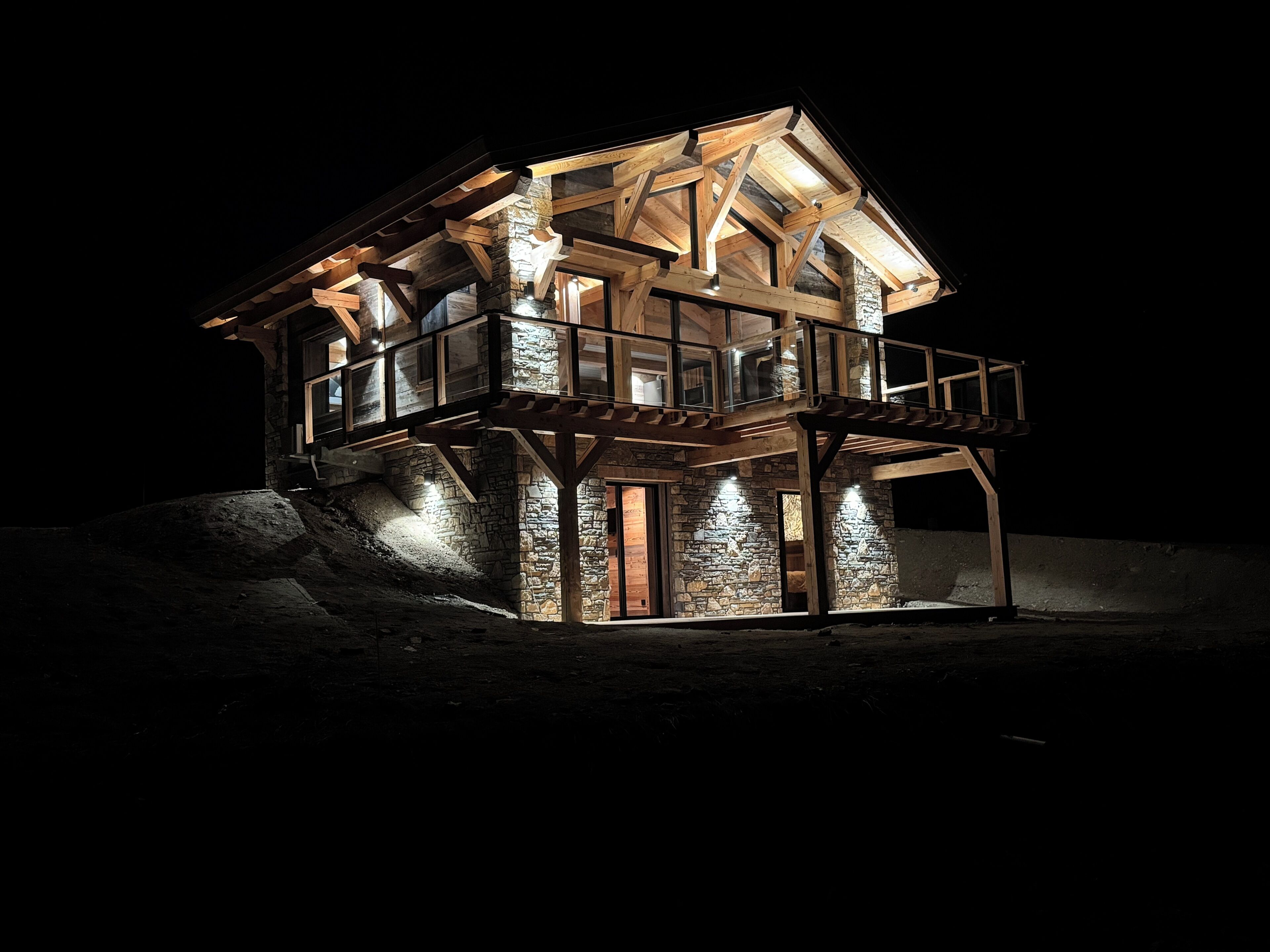 A multi-story wooden house with a balcony and stone walls at night.