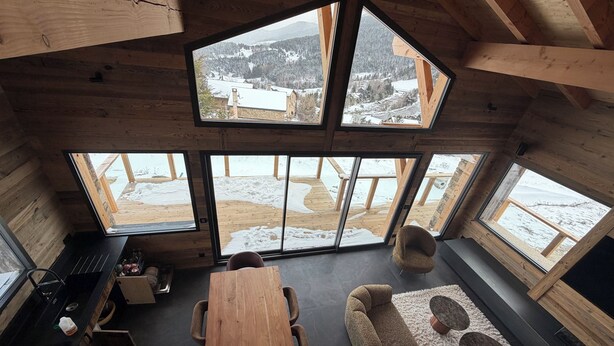 A wooden cabin interior with a dining table, sofa, and large windows offering a snowy mountain view. - Luxury 115-square-meter chalet in a ski resort, ideal for two families (8–10 people) (Saint-Pierre-dels-Forcats)