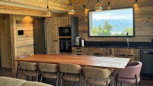 A wooden interior with a large dining table, chairs, a kitchen area, and a window with a mountain view. - Luxury 115-square-meter chalet in a ski resort, ideal for two families (8–10 people) (Saint-Pierre-dels-Forcats)