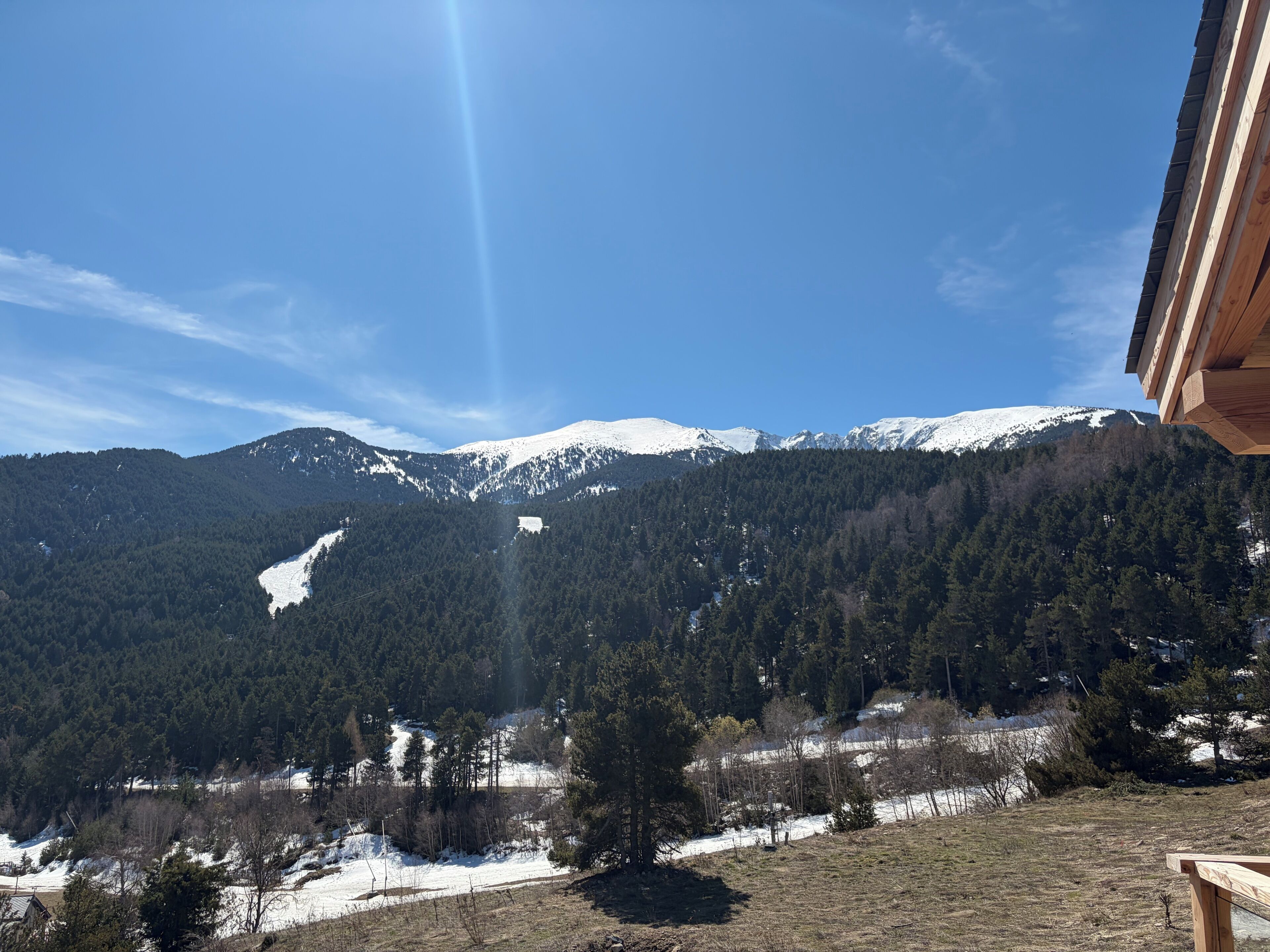 A mountain landscape with snow-covered peaks, a forested valley, and a building with a sloped roof.