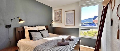 A modern bedroom with a bed, bedside table, lamp, artwork, and a view of the outside.