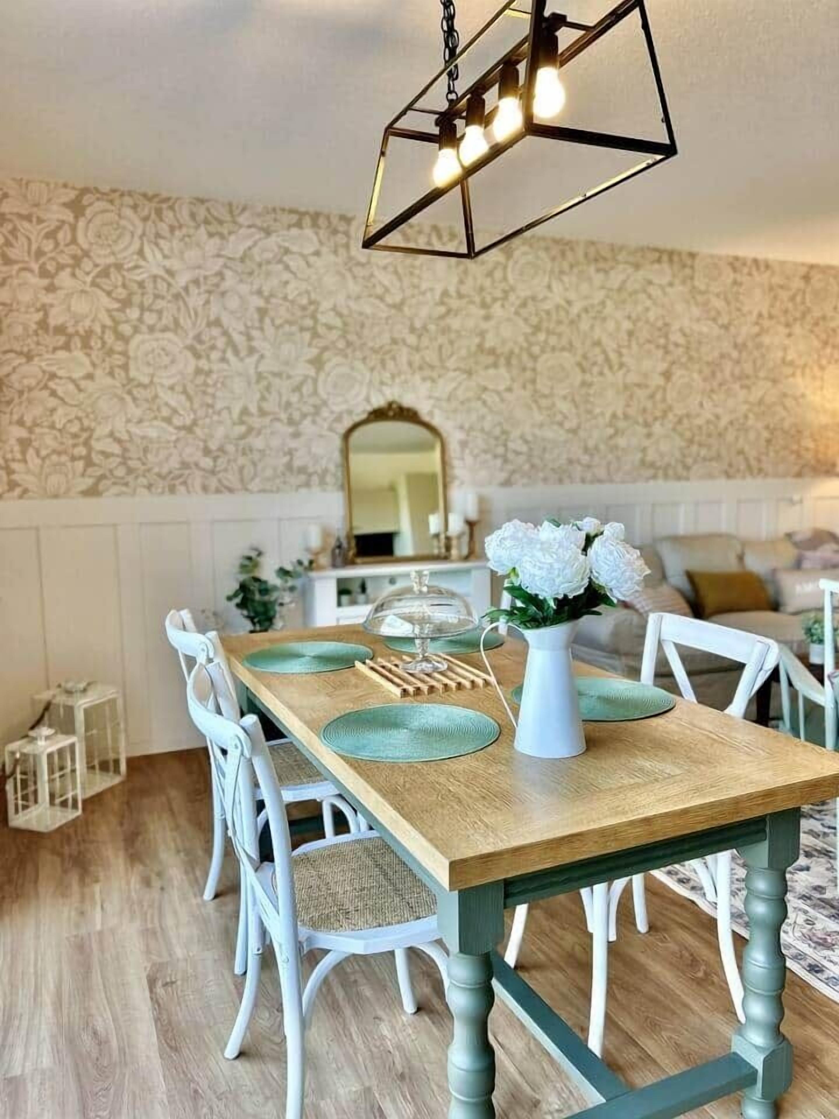 Un comedor con mesa de madera, sillas blancas, lámpara colgante y papel tapiz con estampado floral.