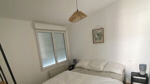 A bedroom with a bed, a bedside table, a lamp, a window with blinds, and a radiator. - Air-conditioned house in an ideal location (Montpellier-de-Médillan)