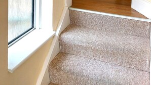Une pièce avec une fenêtre, un lit, un ventilateur sur pied et un escalier recouvert de moquette.