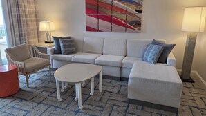 Une chambre d’hôtel moderne avec un canapé gris, une table basse ronde, un fauteuil blanc et un tapis à motifs.