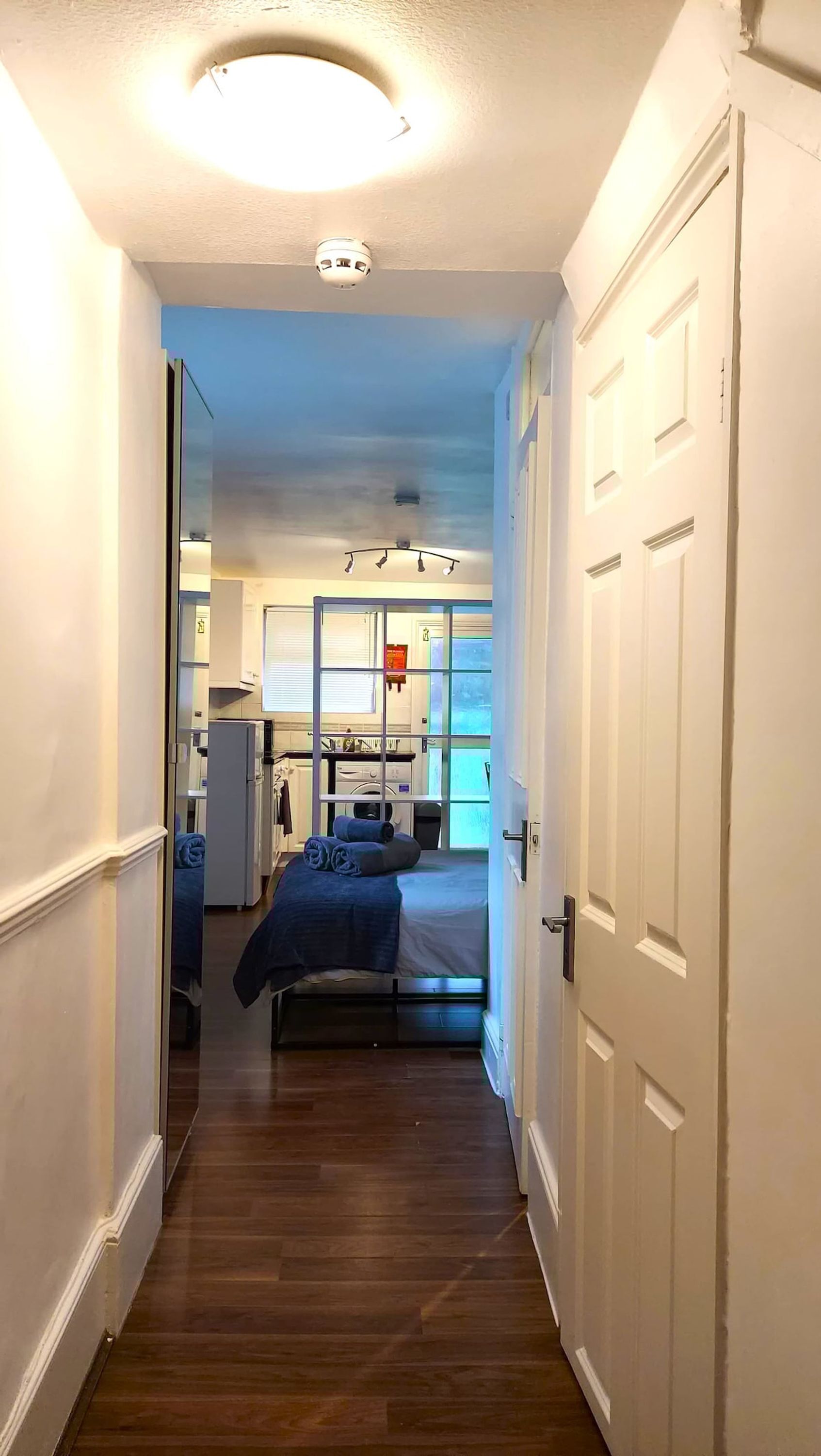 A narrow hallway leading to a bedroom with a bed, a window, and a visible laundry area.