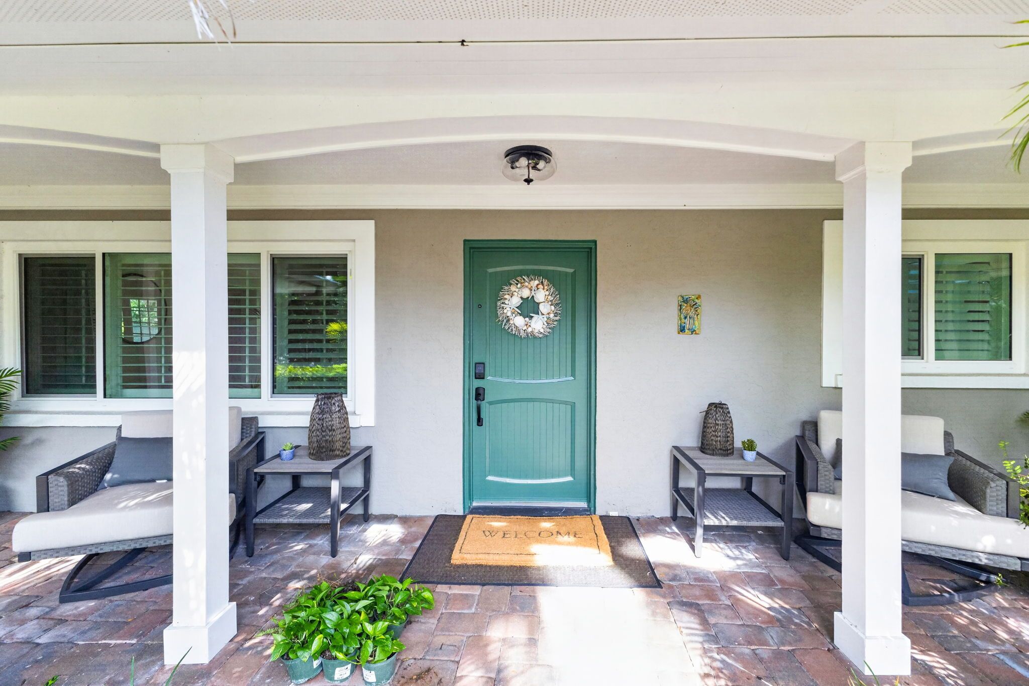 A covered porch with a green door, two chairs, a welcome mat, and potted plants.