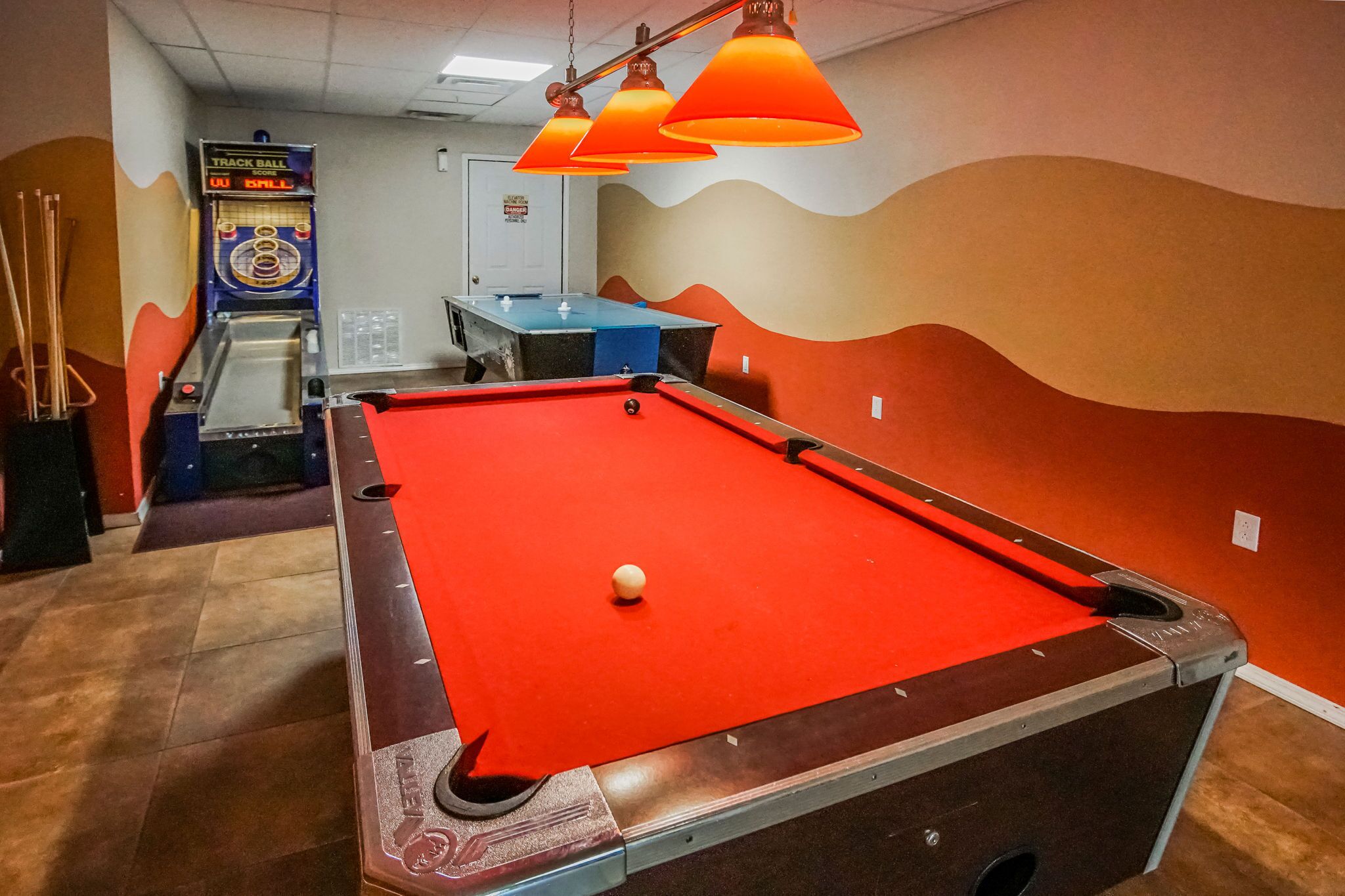Une table de billard recouverte d’un drap de feutre rouge, une boule blanche et des luminaires suspendus de couleur orange.