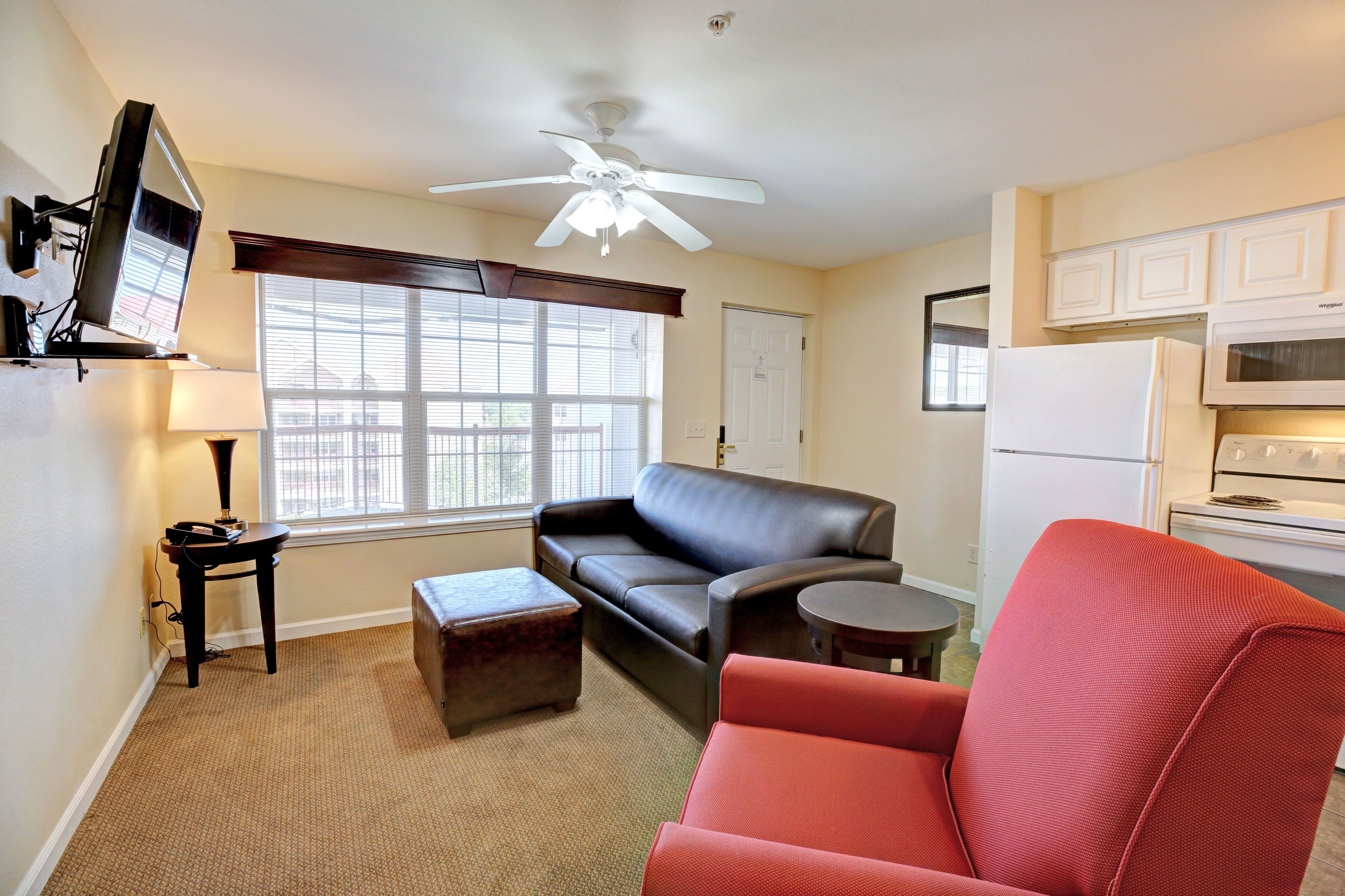 A living room with a sofa, ottoman, and a red chair. There is a television, a ceiling fan, and a window with blinds.
