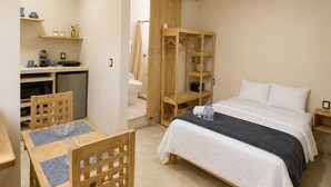 Una habitación de hotel compacta con cama, una pequeña cocina americana, una mesa de comedor de madera con sillas y un armario.