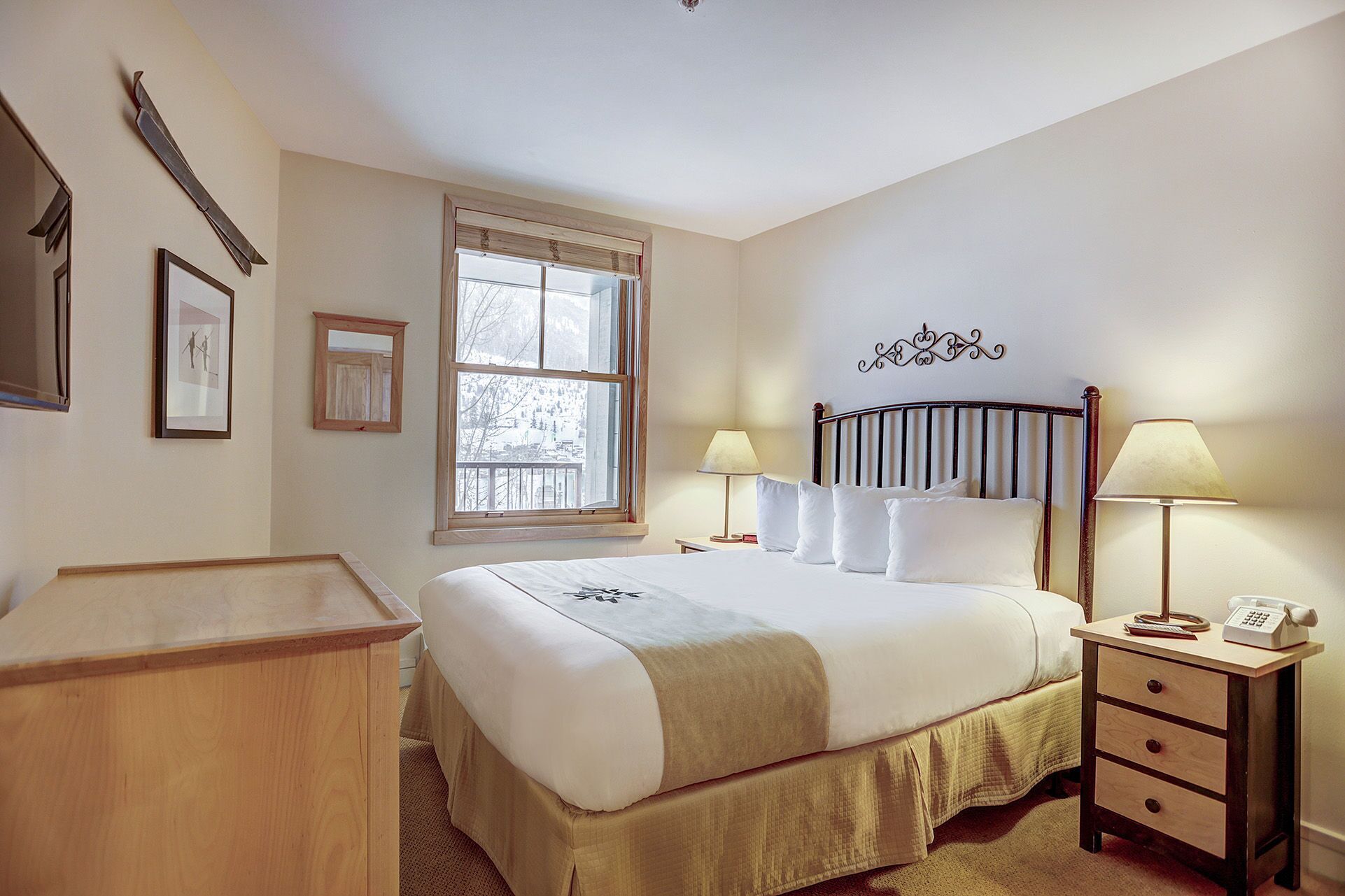 A neatly arranged bedroom with a bed, bedside lamps, a wooden nightstand, a desk, and a window with a view of trees.