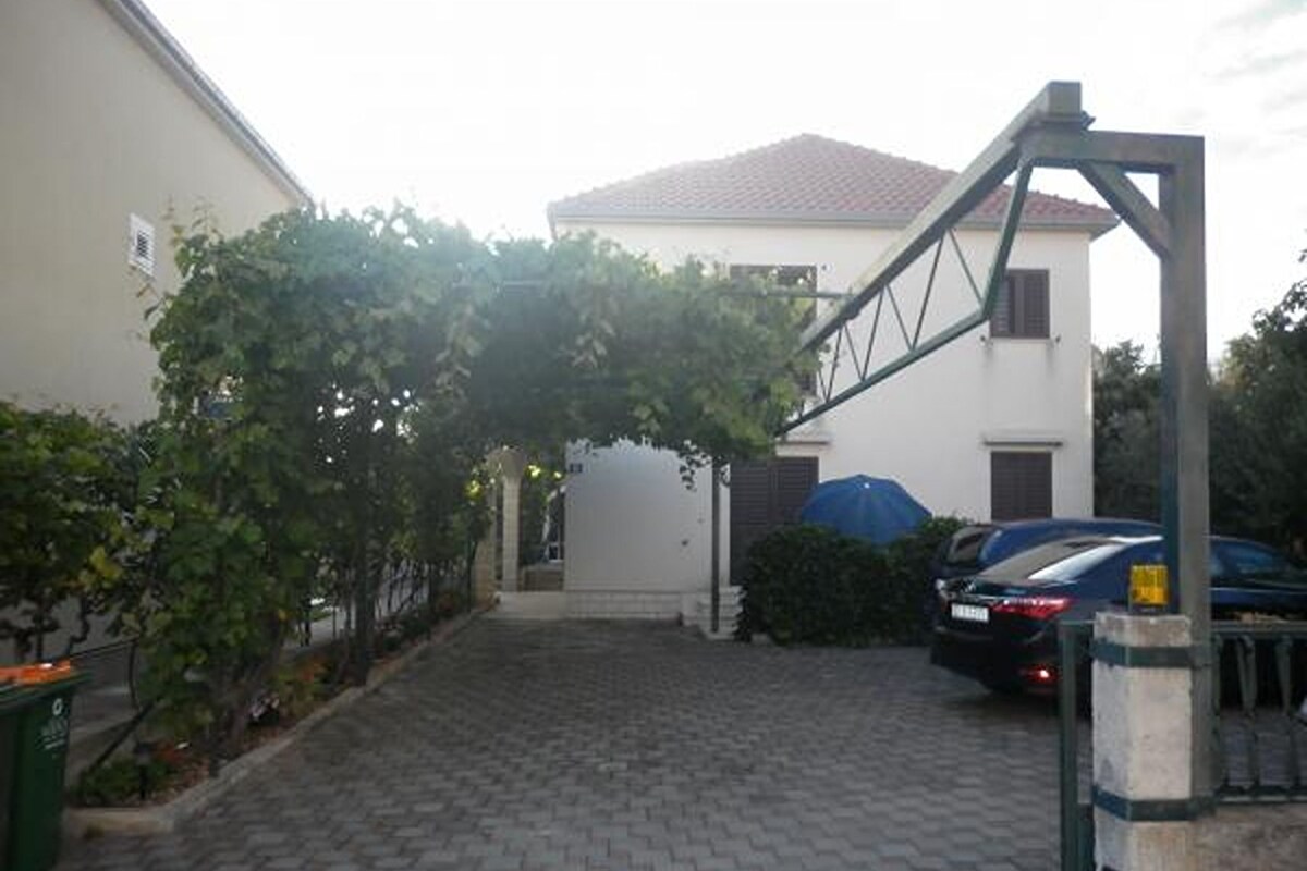 A paved driveway leading to a house with a covered entrance, a car parked in the driveway, and a green trash bin to the left.