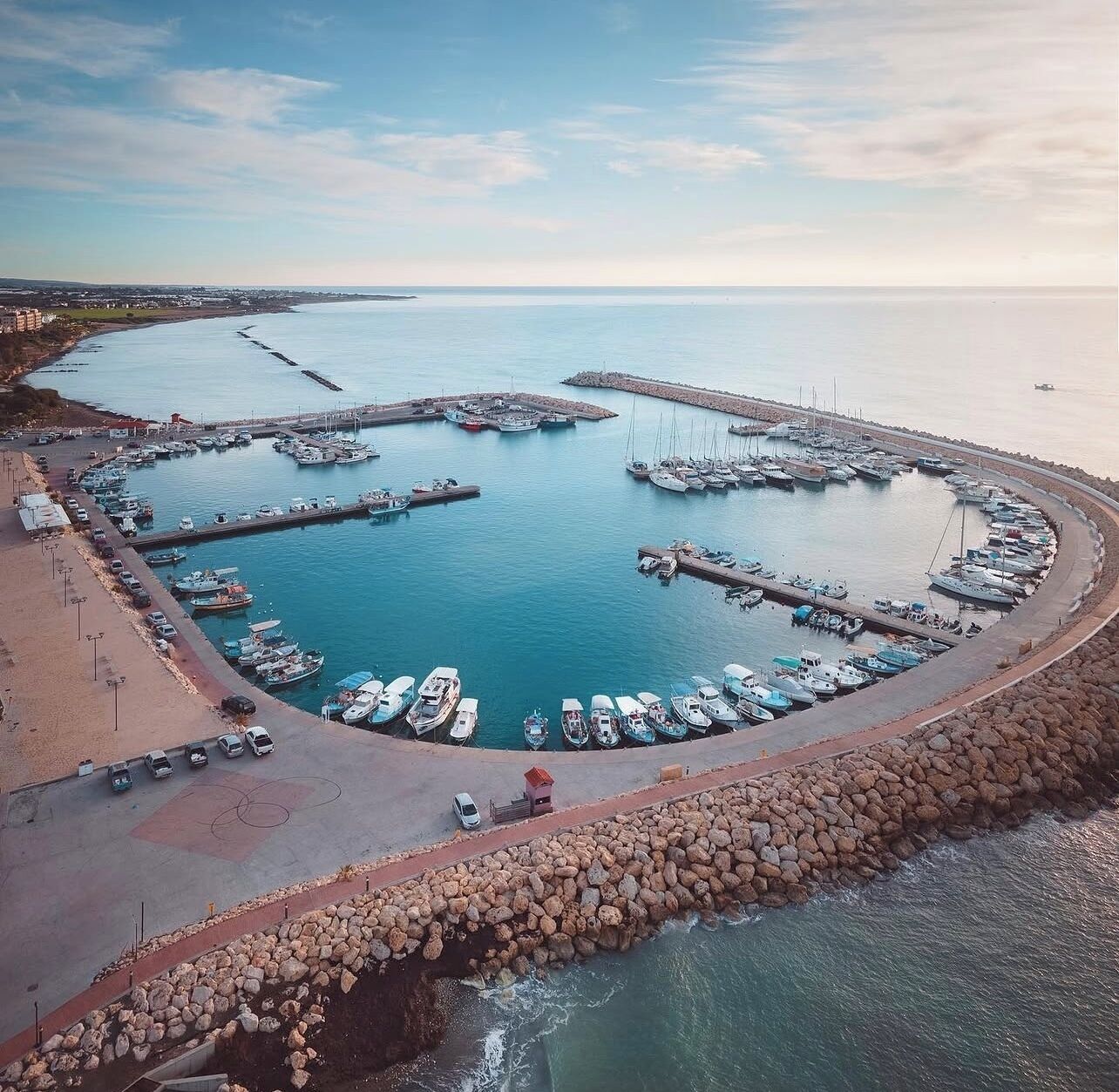A marina with boats docked, a rocky breakwater, and a clear sky.