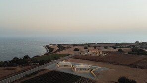 An aerial view of a coastal property with a road leading to a building complex, fields, and a coastline.