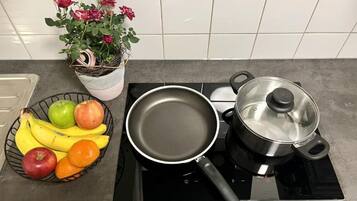 Une cuisine avec une casserole sur une plaque de cuisson à induction, une poêle, un panier de fruits et une plante en pot.
