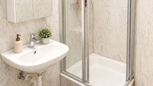 Un baño con ducha de vidrio, un lavamanos blanco con un espejo encima y una pequeña planta en maceta sobre la encimera.