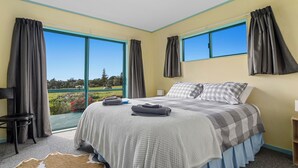 A bedroom with a large bed, two windows, a chair, and a view of a green landscape. - Little Kowhai - Te Kaha Holiday Home (Te Kaha)