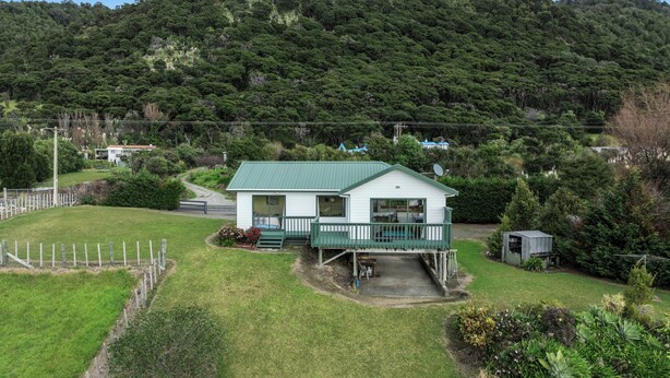 A small house with a green roof and white walls, surrounded by a fenced yard and lush greenery. - Little Kowhai - Te Kaha Holiday Home (Te Kaha)