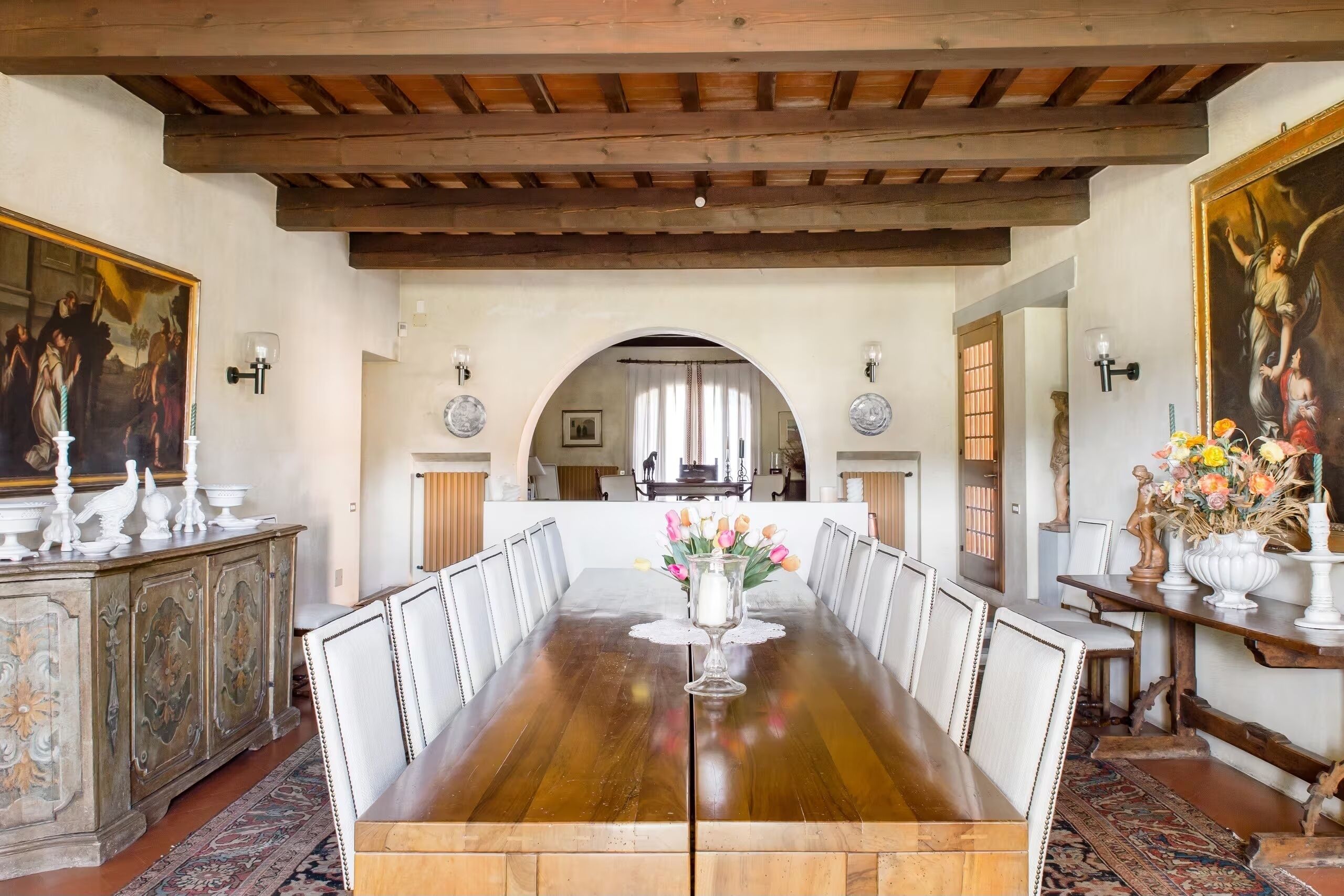 Una sala da pranzo con un lungo tavolo di legno, sedie bianche, un vaso di fiori e un quadro appeso al muro.