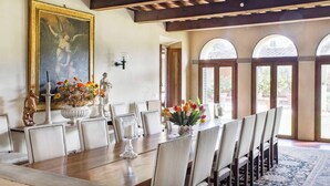 Une salle à manger avec une longue table, des chaises, un vase de fleurs, un tableau et des fenêtres en arc de cercle.