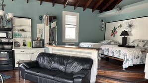 Una habitación acogedora con un sofá de cuero negro, una cama con ropa de cama floral, una escalera de madera y una zona de cocina con microondas.