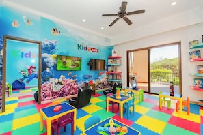 Un espace de jeux pour enfants avec un sol coloré, un ventilateur de plafond et un écran de télévision.