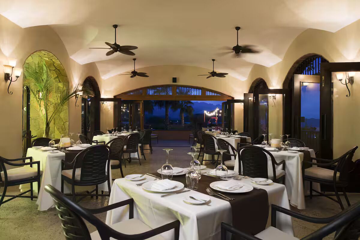 Um restaurante com mesas preparadas para refeições, ventiladores de teto e vista para o céu noturno.
