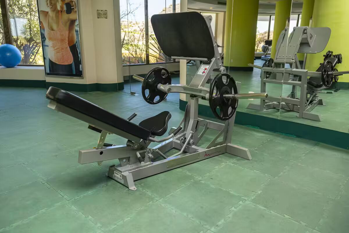 Et fitnesscenter med træningsudstyr, herunder bænkpres og vægtmaskiner, samt et stort spejl og vinduer.