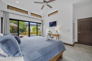 Une chambre dotée d’un grand lit, d’un bureau avec une chaise, d’un ventilateur de plafond et offrant une vue sur une piscine et un espace verdoyant.