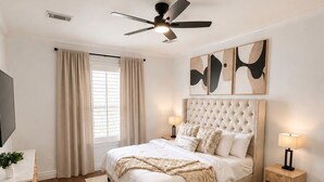 Ein Schlafzimmer mit einem Bett, einem Schreibtisch, einem Sessel und einem Deckenventilator, außerdem Kunstwerken an der Wand.