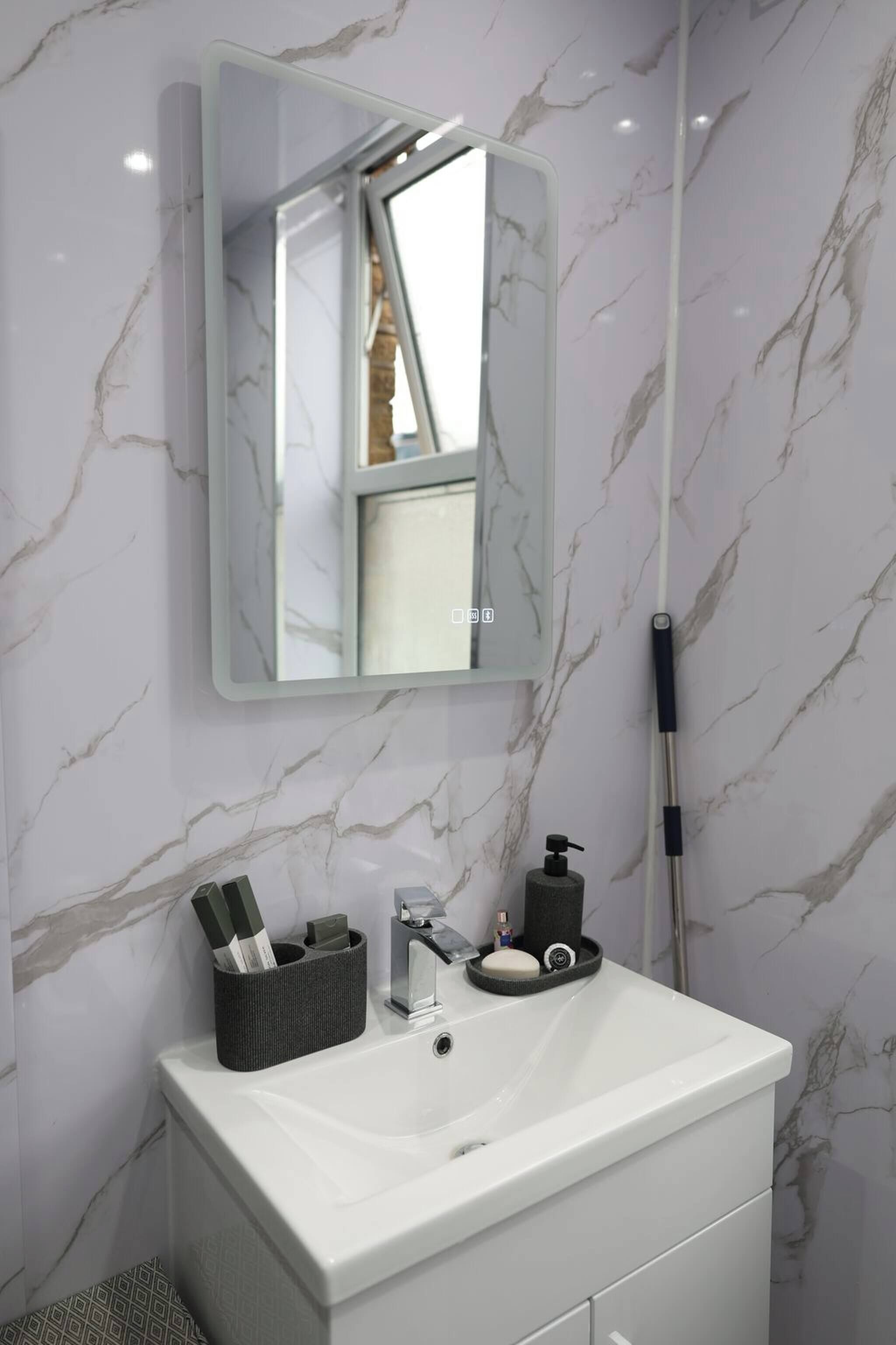 Une salle de bain avec un lavabo blanc, un miroir, un porte-brosse à dents, un distributeur de savon et une serpillière.