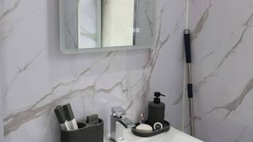 Une salle de bain avec un lavabo blanc, un miroir, un porte-brosse à dents, un distributeur de savon et une serpillière.