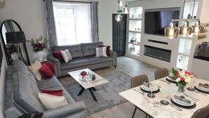 Un salon moderne avec un canapé gris, une table à manger pour quatre personnes, un téléviseur à écran plat et une cheminée.