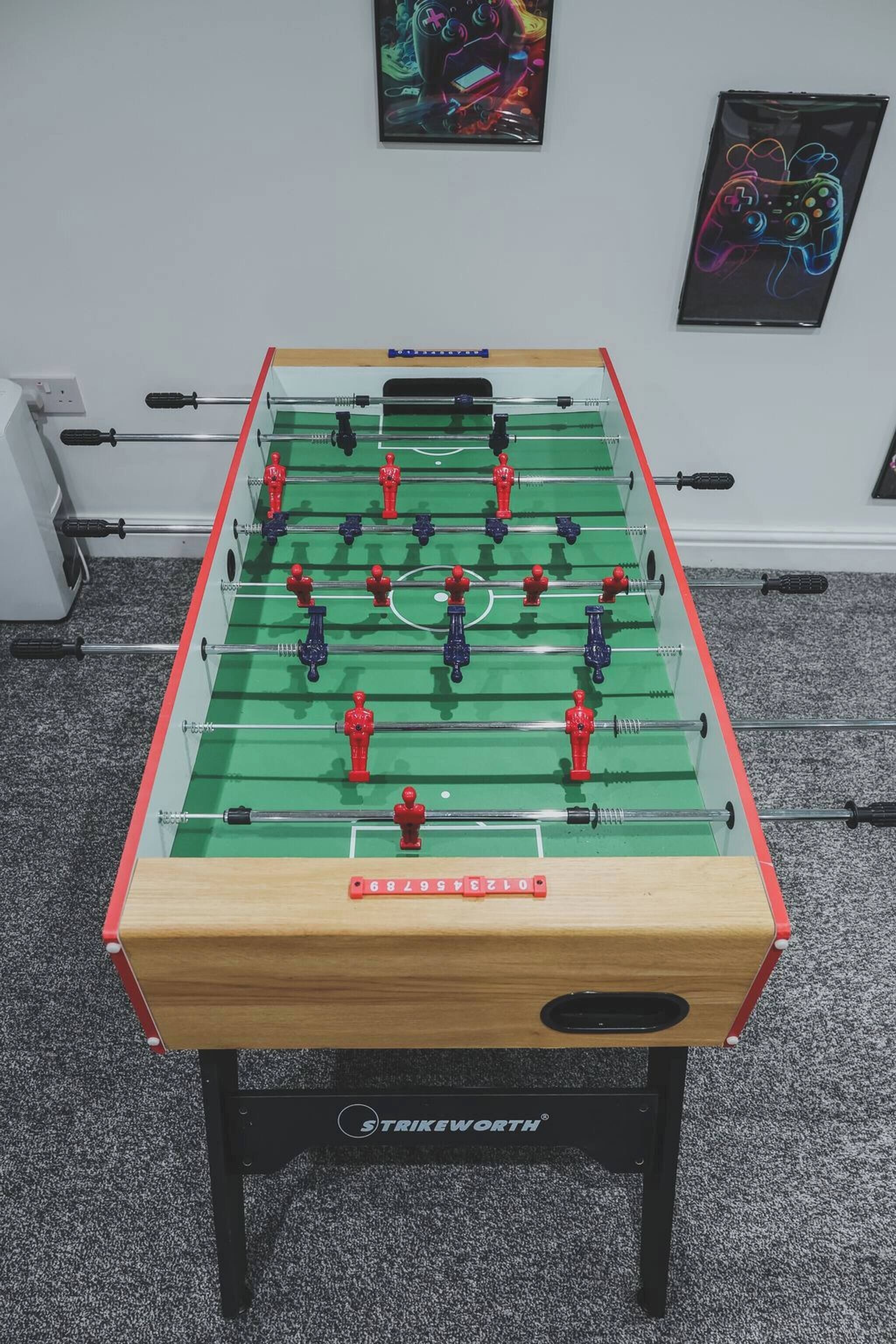 Un tavolo da calcio con base in legno e aste metalliche, posto di fronte a una parete con due quadri incorniciati.