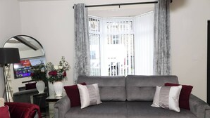 Un canapé gris avec deux coussins, un vase avec des fleurs et un téléviseur posé sur un meuble.