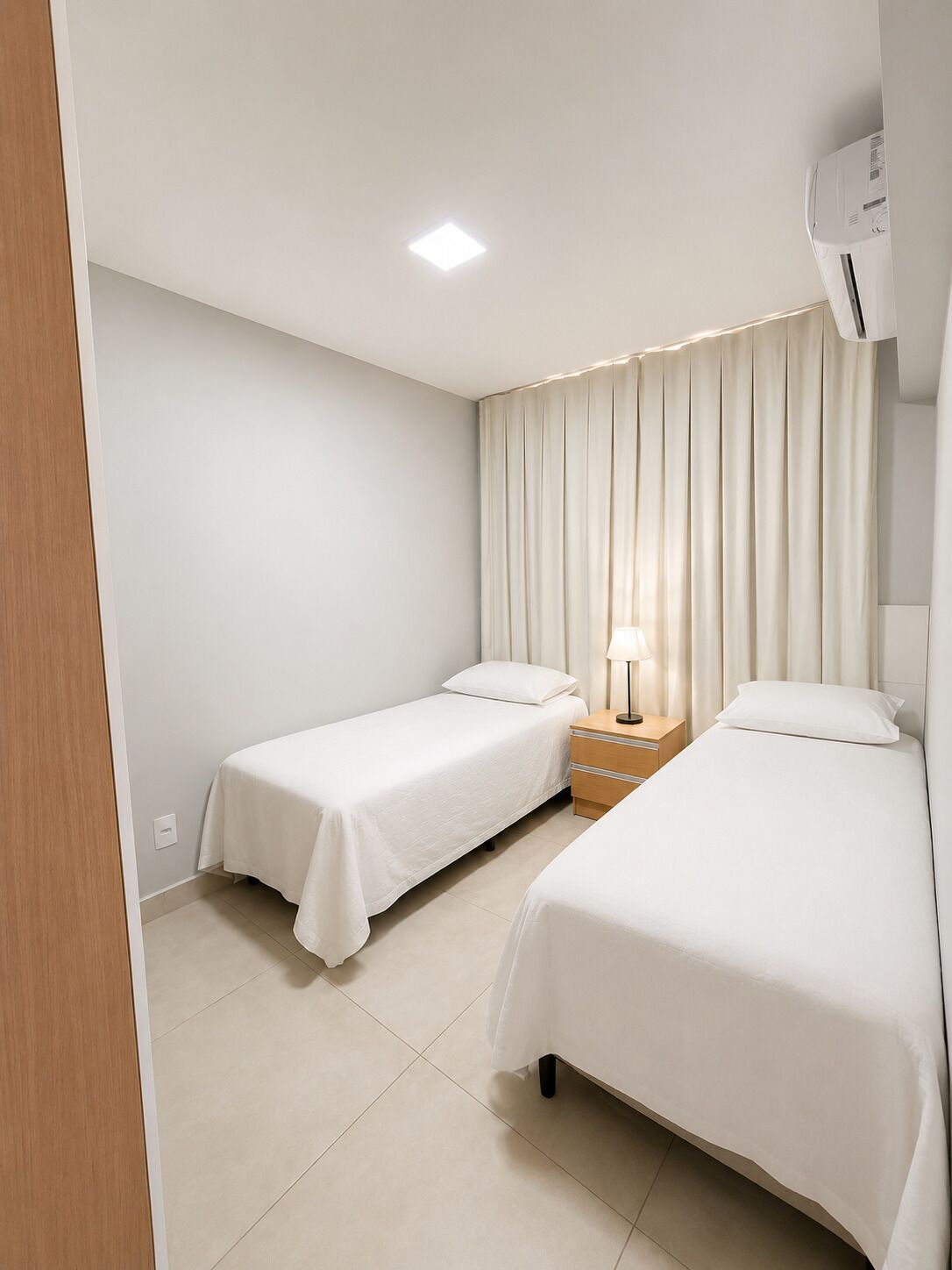 Une chambre d’hôtel avec deux lits simples, une table de chevet avec une lampe, un climatiseur fixé au mur et un luminaire au plafond.