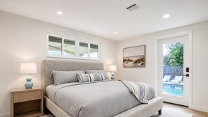 A bedroom with a large bed, two bedside tables, a window, a door to a pool area, and a painting on the wall.