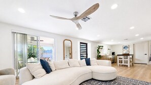 A modern living room with a large L-shaped sectional sofa, a ceiling fan, and a kitchen area in the background.