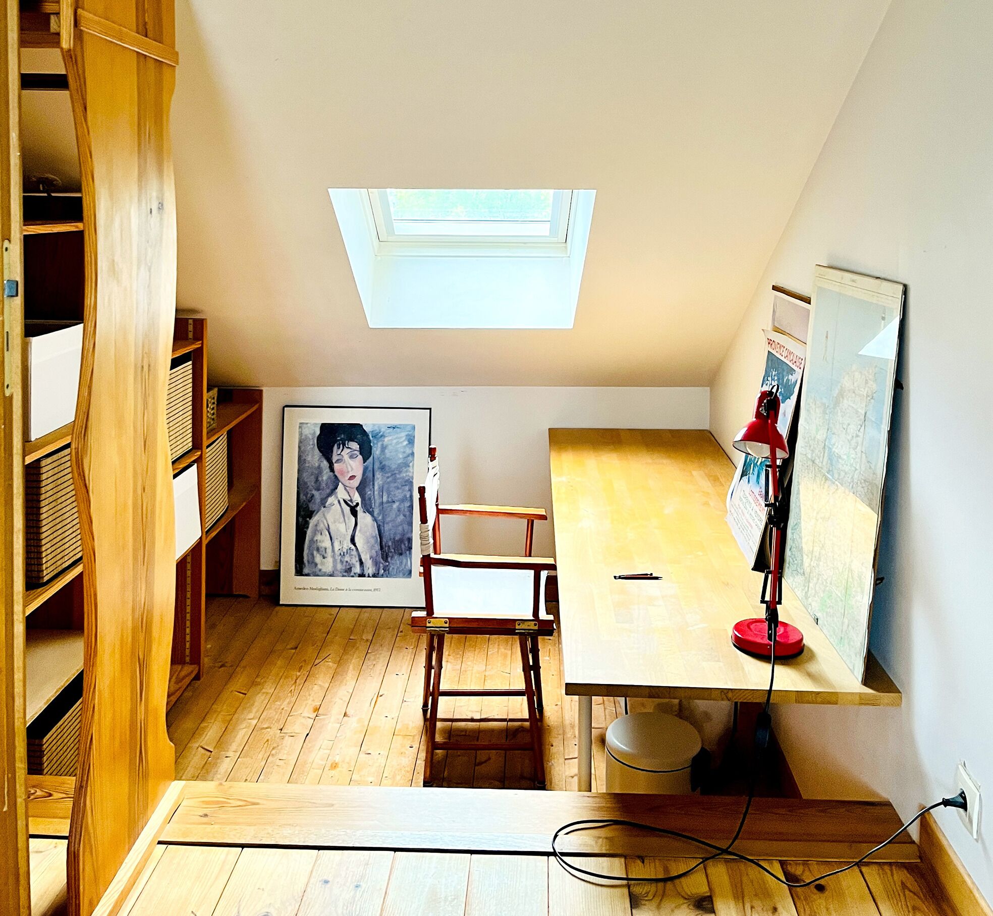 Une pièce avec un plafond en pente, un puits de lumière, un bureau en bois, une chaise, un tableau encadré et une lampe rouge.