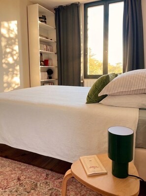 Ein Schlafzimmer mit einem Bett, einem Nachttisch mit einem Buch und einer grünen Tasse, einem Bücherregal und einem Fenster mit Vorhängen.