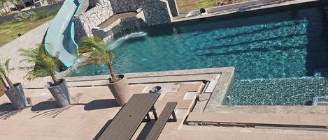 A swimming pool with a slide, surrounded by a stone wall and a picnic table.