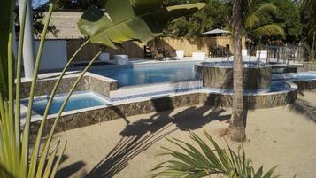 A swimming pool with a stone edge and blue mosaic tiles, surrounded by palm trees and a wooden fence.