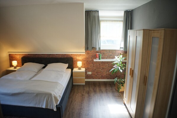 Une chambre à coucher avec un lit, des tables de chevet, une commode, une armoire, un mur en briques, une fenêtre avec des rideaux et une plante en pot.