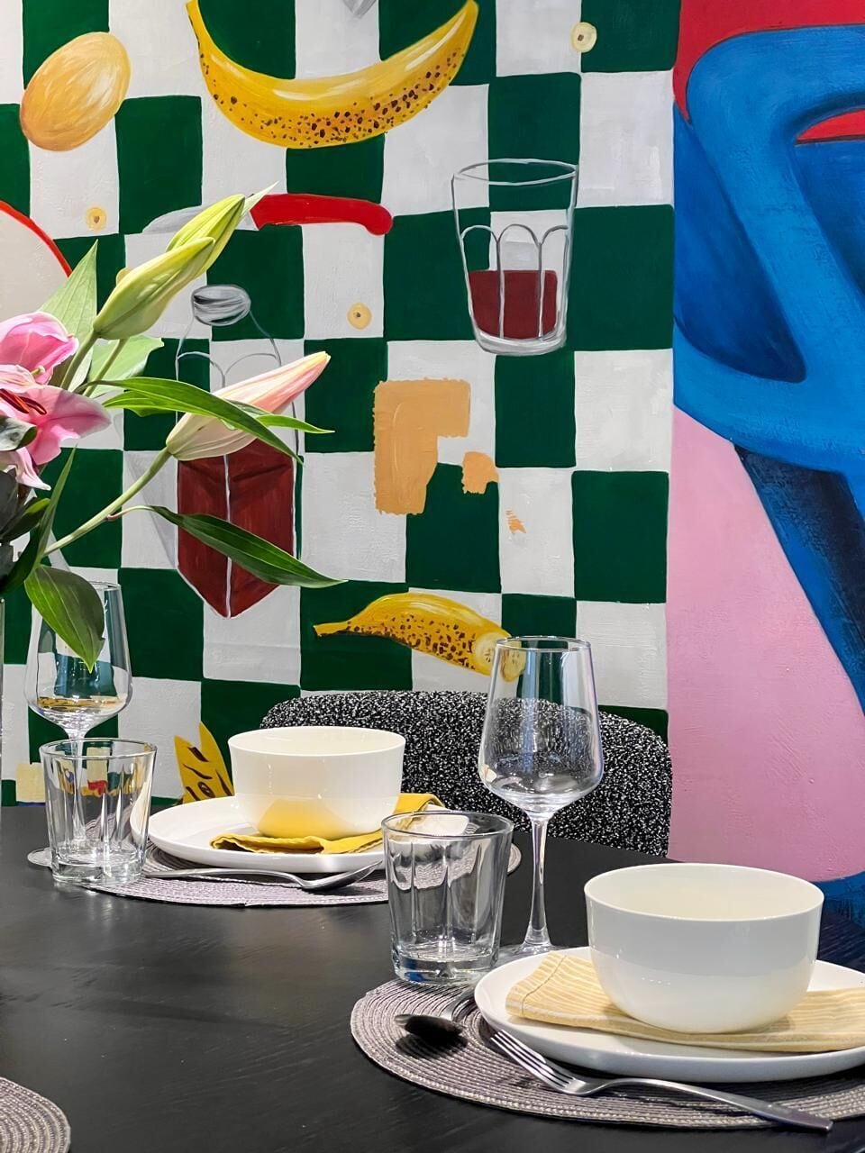 Une table à manger dressée avec des assiettes, des verres et des couverts, et une peinture abstraite aux couleurs vives en arrière-plan.