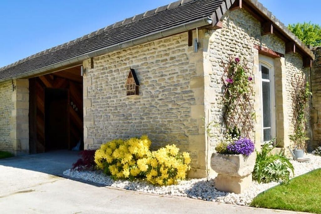 Un bâtiment en pierre doté d’une entrée couverte, d’une petite fenêtre et d’un jardon contenant des fleurs jaunes et violettes.