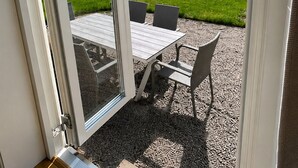 Een terras met tafel en stoelen, een glazen deur en uitzicht op een gazon met bomen.