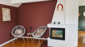 Een kamer met een open haard, twee witte rieten stoelen, een klein rond tafeltje en een wanddecoratie van een blad in een lijst.
