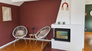 Habitació amb una llar de foc, dues cadires blanques de vimetla, una taula petita rodona i una obra d'art amb fulles emmarcada a la paret.